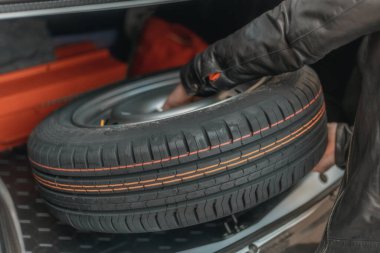 Yedek lastik için bagaja uzanan bir insan eli fotoğrafı. Parlak renkli çizgileri olan yeni bir lastik. Yolda lastik değiştirmek, seyahate hazırlanmak, ciddi bir olay. Araba bakımı.