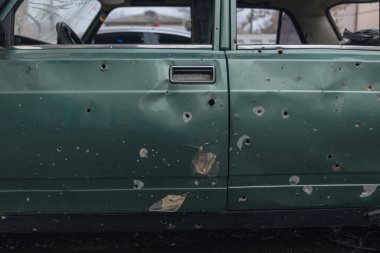 A close-up of a civilian car door riddled with numerous shrapnel and bullet holes. Damage to the paintwork and traces of metal at the impact sites are visible.