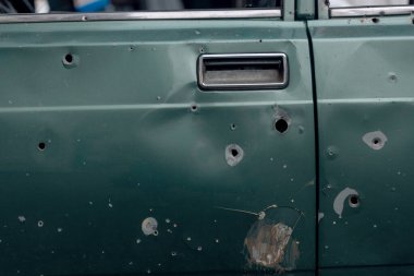 A close-up of a civilian car door riddled with numerous shrapnel and bullet holes. Damage to the paintwork and traces of metal at the impact sites are visible.