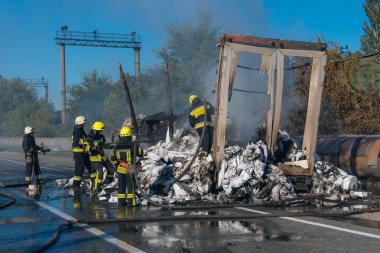 Koruyucu giysili beş itfaiyeciden oluşan bir ekip, büyük bir yangını söndürdükten sonra ıslak bir yolda durup olay yerini inceliyor. Kalın buhar ve duman, yanan enkazdan yükseliyor..