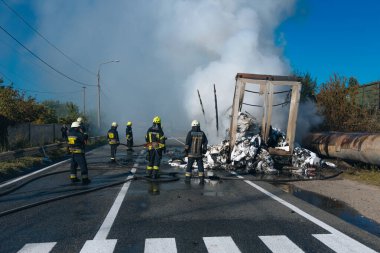 Koruyucu giysili beş itfaiyeciden oluşan bir ekip, büyük bir yangını söndürdükten sonra ıslak bir yolda durup olay yerini inceliyor. Kalın buhar ve duman, yanan enkazdan yükseliyor..