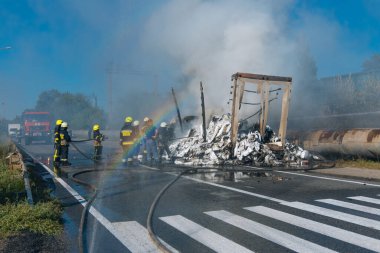 Koruyucu giysili beş itfaiyeciden oluşan bir ekip, büyük bir yangını söndürdükten sonra ıslak bir yolda durup olay yerini inceliyor. Kalın buhar ve duman, yanan enkazdan yükseliyor..