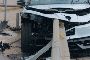 Ciddi bir trafik kazasının ardından ağır hasarlı beyaz bir araba metal korkuluklara çarptı. Arabanın ön tarafı parçalanmış ve enkaz parçaları asfalta saçılmış..
