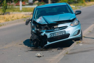Ciddi bir kazadan sonra hurdaya dönmüş mavi bir araba. Ön manzara, çarpışmadan kaynaklanan ağır hasar görmüş kaput, tampon ve motoru kaldırımda enkaz ve sıvı lekeleri gösteriyor..