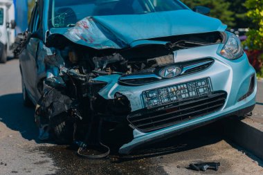 Ciddi bir kazadan sonra hurdaya dönmüş mavi bir araba. Ön manzara, çarpışmadan kaynaklanan ağır hasar görmüş kaput, tampon ve motoru kaldırımda enkaz ve sıvı lekeleri gösteriyor..