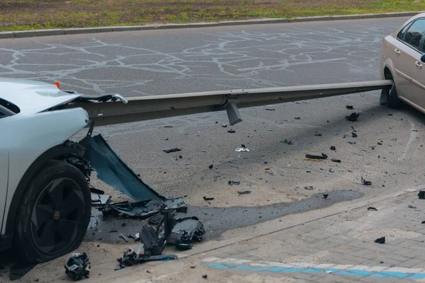 Ciddi bir trafik kazasının ardından ağır hasarlı beyaz bir araba metal korkuluklara çarptı. Arabanın ön tarafı parçalanmış ve enkaz parçaları asfalta saçılmış..