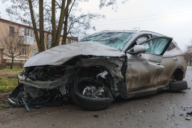 Şiddetli bir kazadan sonra, bir şehir caddesinde meydana gelen çarpışmadan sonra ön yüzünde feci hasar olan gri bir SUV. Kaput buruşmuş, lastik yerinden çıkmış, ön cam kırılmış ve kapı açık ve deforme olmuş..
