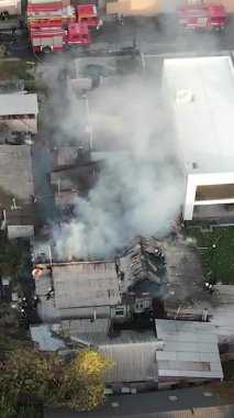 Bir yerleşim bölgesinde büyük bir yangınla mücadele: özel bir evin kömürleşmiş çatısında çalışan itfaiyecilerin havadan görünüşü, mahallenin üzerine yoğun duman yükseliyor. Kurtarma ekipleri ve itfaiye araçları olay yerinde..