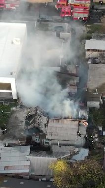 Bir yerleşim bölgesinde büyük bir yangınla mücadele: özel bir evin kömürleşmiş çatısında çalışan itfaiyecilerin havadan görünüşü, mahallenin üzerine yoğun duman yükseliyor. Kurtarma ekipleri ve itfaiye araçları olay yerinde..