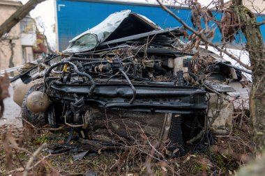 Ciddi bir trafik kazasının ardından: tamamen tahrip olmuş bir ön ucu olan beyaz bir yolcu arabası, şehir caddesinin kenarında bir ağaca çarptı. Açık motor bölmesi, buruşuk kaput, kırık ön cam ve deforme olmuş kapılar görülebilir durumda.