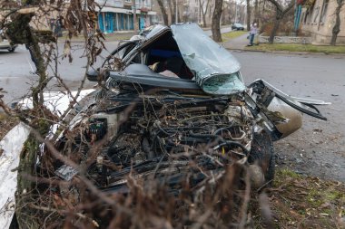 Ciddi bir trafik kazasının ardından: tamamen tahrip olmuş bir ön ucu olan beyaz bir yolcu arabası, şehir caddesinin kenarında bir ağaca çarptı. Açık motor bölmesi, buruşuk kaput, kırık ön cam ve deforme olmuş kapılar görülebilir durumda.