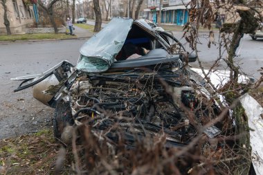 Ciddi bir trafik kazasının ardından: tamamen tahrip olmuş bir ön ucu olan beyaz bir yolcu arabası, şehir caddesinin kenarında bir ağaca çarptı. Açık motor bölmesi, buruşuk kaput, kırık ön cam ve deforme olmuş kapılar görülebilir durumda.