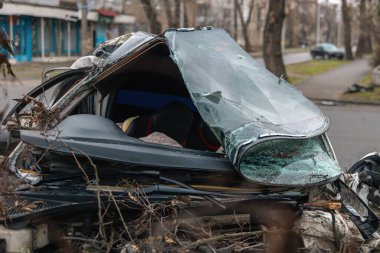 Ciddi bir trafik kazasının ardından: tamamen tahrip olmuş bir ön ucu olan beyaz bir yolcu arabası, şehir caddesinin kenarında bir ağaca çarptı. Açık motor bölmesi, buruşuk kaput, kırık ön cam ve deforme olmuş kapılar görülebilir durumda.