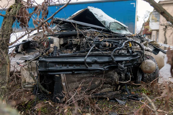 Ciddi bir trafik kazasının ardından: tamamen tahrip olmuş bir ön ucu olan beyaz bir yolcu arabası, şehir caddesinin kenarında bir ağaca çarptı. Açık motor bölmesi, buruşuk kaput, kırık ön cam ve deforme olmuş kapılar görülebilir durumda.