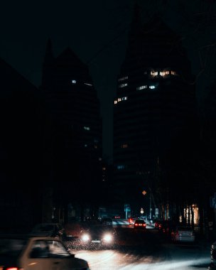 İki modern, ikiz kule tarzı gökdelenlerin dramatik gece manzarası. Ön planda, hareket halindeki arabalarla karla kaplı bir şehir caddesi, farları derin gölgeler ve karanlık gökyüzüne karşı canlı aksanlar yaratıyor..