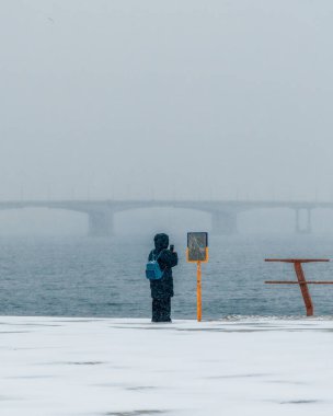 Yoğun kar yağışı sırasında cep telefonuyla fotoğraf çeken bir adam. Soğuk suyun üzerindeki sislerin içinde kaybolan köprünün görüntüsü modern bir kış yürüyüşünün minimalist ve sinematik bir görüntüsünü ve dijital olarak yakalanan bir anı yaratır..