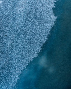 Donmuş su yüzeyinin ince bir buz tabakası ya da kar taneleri ile kaplı soyut bir görüntüsü. Görüntü, buzun derin mavisi ile yüzeyindeki beyaz kristaller arasındaki zıtlığı vurgular..