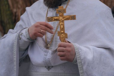 Kış ayini sırasında elinde altın ayin haçı tutan bir Ortodoks rahibinin yakın çekimi. Rahip gümüş işlemeli geleneksel beyaz cübbeler giyiyor ve zincirli bir göğüs kafesi takıyor..