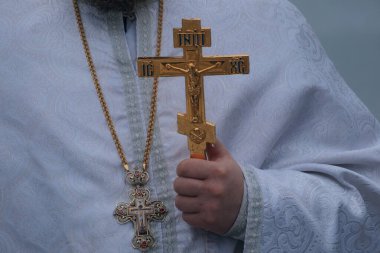 Kış ayini sırasında elinde altın ayin haçı tutan bir Ortodoks rahibinin yakın çekimi. Rahip gümüş işlemeli geleneksel beyaz cübbeler giyiyor ve zincirli bir göğüs kafesi takıyor..