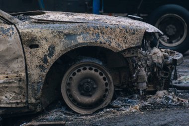 Bombardımandan kalan kül ve çamurla kaplı harap olmuş sivil bir arabanın yakın çekimi. Patlak bir lastik ve yanmış bir çamurluk askeri harekatın trajik ve yıkıcı sonuçlarını sembolize eder..