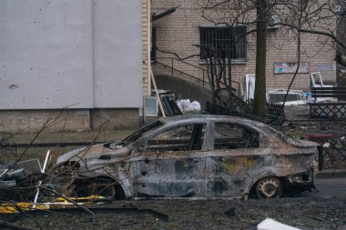 Hasarlı bir apartmanın önünde tamamen yanmış bir araba. Şarapnel ve ateş kalıntıları, binanın gövdesinde ve gölgesinde kaldı. Çatışma sırasında sivil işçilerin yıkımına dair çarpıcı kanıtlar.