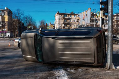 Bir araba kazasından sonra şehrin kavşağında altın rengi bir SUV devrilmiş. Olay yeri, güneşli bir kış gününde arka planda trafik ışıkları, yol işaretleri ve konut binalarıyla kaldırımdaki hasarlı aracı yakalıyor.
