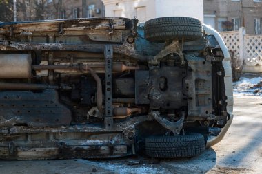 Trafik kazasından sonra devrilmiş bir SUV 'un şasisi ve ön süspansiyonunun detaylı bir orta boy çekimi. Görüntü, şehrin caddesinde bulunan aracın karmaşık mekanik bileşenleri, şasisi ve kirli gövde panellerini gösteriyor.