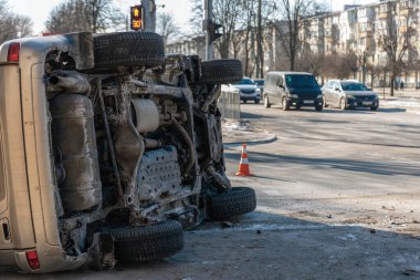 Trafik kazasından sonra devrilen bir cipin geniş açılı kentsel görüntüsü. Görüntü aracın şehir caddesinde trafiği, yayaları ve arka planda trafik ışığı olan bir araç ve vücut hasarını gösteriyor.