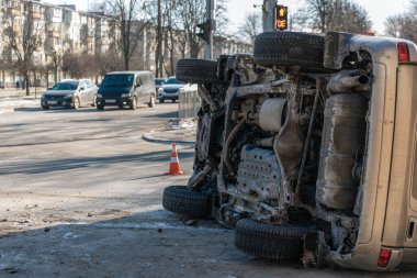 Trafik kazasından sonra devrilen bir cipin geniş açılı kentsel görüntüsü. Görüntü aracın şehir caddesinde trafiği, yayaları ve arka planda trafik ışığı olan bir araç ve vücut hasarını gösteriyor.