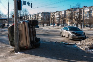 Kış boyunca şehirde meydana gelen bir trafik kazası sahnesinin sokak seviyesinde geniş bir görüntüsü. Önü hasarlı gümüş bir sedan devrilmiş altın bir cipin yanında duruyor. Bir kavşakta elektrik direğine çarpmış.