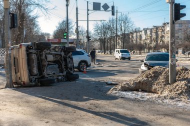 Güneşli bir kış gününde şehir kavşağında meydana gelen trafik kazasının geniş şehir görüntüsü. Önü hasarlı gümüş renkli bir sedan yakınlarda dururken, yan yatmış bir altın SUV elektrik direğine çarpıyor. Yayalar ve diğer araçlar
