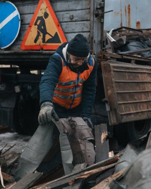 Turuncu yansıtıcı yelek ve siyah şapka enkazı temizleyen bir kamu görevlisi. Yol tabelası olan bir kamyonun önündeki moloz yığınından buruşmuş bir metal parçası kaldırıyor..