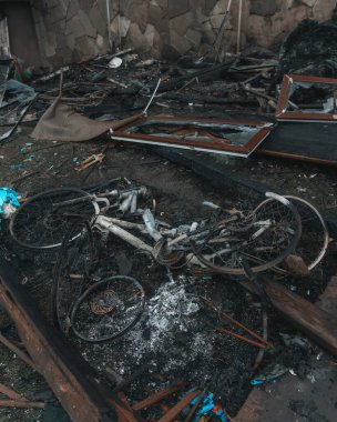 Tamamen yanmış bisiklet iskeleti ve tekerlekler kül ve yanmış molozların içinde yatıyor. Yakınlarda kırık camlı kırık bir pencere çerçevesi var. Zemin is, kömür ve toprak parçalarıyla kaplı..