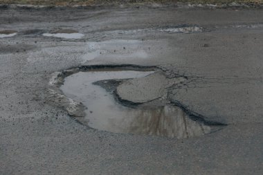 Karanlık bir asfalt yolda büyük, hilal şeklinde bir çukurun detaylı ve yüksek açılı bir görüntüsü. Delik derin ve bulanık suyla doludur. Etrafı çatlamış ve ufalanan asfalt kenarlarıyla doludur. Bu görüntü, kötüleşen...