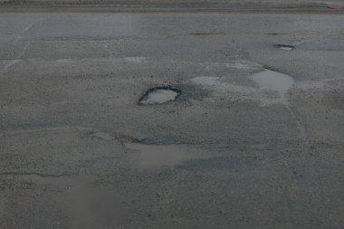 Gri asfalt bir yol yüzeyinin erozyon ve ihmalden ağır hasar görmüş yukarıdan aşağı yakın çekimi. Çeşitli boyutlarda birkaç derin çukur görünür, durgun, bulanık yağmur suyuyla doludur. Yıpranmış asfaltın dokusu pürüzlü ve çatlamış.,