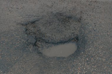 Yıpranmış asfalt bir yolda küçük, derin bir çukurun yakın çekim, tepeden aşağı görüntüsü. Çukur bulanık suyla dolu ve çatlamış, ufalanan kaldırımlarla çevrili. Bu da ciddi altyapı ihmalinin altını çiziyor.
