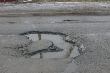 Gri asfalt bir yolda büyük, derin bir çukurun yüksek açılı yakın çekimi. Krater benzeri delik bulanık suyla doludur. Çentikli, ufalanan kenarlar ve merkezde çatlamış bir asfalt adası vardır..