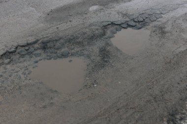 Gri asfalt bir yol yüzeyinin yüksek açılı görüntüsü ileri derecede bozulmayı gösteriyor. Bulanık yağmur suyuyla dolu iki büyük, kalp şeklinde çukur belirgindir. Şiddetli çatlaklar ve parçalanan kaldırımlarla çevrilidir.