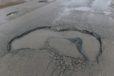 Yüksek açılı bir çekim, çatlak bir asfalt yolda bulanık suyla dolu büyük ve düzensiz şekilli bir çukuru tasvir eder. Çevresindeki asfalt derin çatlaklar ve çökmüş kenarlarla ağır bir şekilde yıpranmış. Bu da önemli altyapı ihmalini gösteriyor.