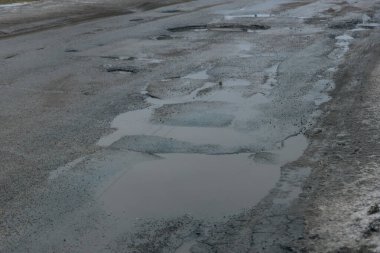 Uzun bir asfalt yolun perspektif çekimi bir dizi derin, su dolu çukurlar tarafından ağır hasar gördü. Parçalanan kaldırım ve büyük su birikintileri araçlar için tehlikeli bir yol oluşturuyor, kasvetli, bulutlu gökyüzünü yansıtıyor.