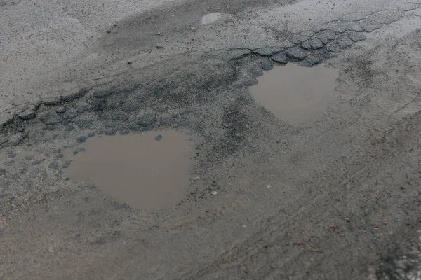 Gri asfalt bir yol yüzeyinin yüksek açılı görüntüsü ileri derecede bozulmayı gösteriyor. Bulanık yağmur suyuyla dolu iki büyük, kalp şeklinde çukur belirgindir. Şiddetli çatlaklar ve parçalanan kaldırımlarla çevrilidir.