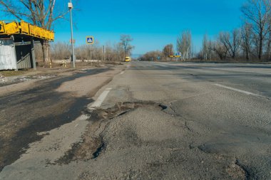 Düşük açılı perspektif çekimi, geniş, güneşli asfalt bir otoyolun ön plandaki büyük ve derin bir çukurdan ciddi şekilde zarar gördüğünü gösteriyor. Yol çıplak kış ağaçları, sarı bir otobüs durağı ve açık mavi gökyüzü ile çevrili.