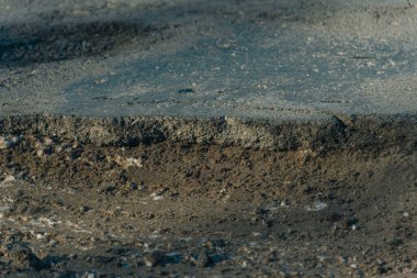 Dar, yatay bir yakın plan, ağır hasar görmüş asfalt bir yolun kesitini yakalar. Çekim, gevşek toprak, kum ve küçük taşlardan oluşan aşınmış bir temelin üzerinde duran kalın, pürüzlü bir kaldırım tabakasını vurguluyor..