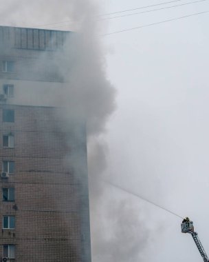 Çok katlı gri tuğlalı bir bina, üst katlardan yükselen kalın gri duman. Sağ alt köşede, bir itfaiyeci Magirus döner merdiveninin sepetinde görünür, ateşin kaynağına doğru bir su akıntısı hedefler.