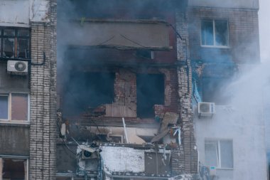 Yüksek katlı bir apartmanın aşırı yakın çekimi bir yangın ya da patlamadan kaynaklanan şiddetli yapısal yıkımı gösteriyor. Korunmasız tuğla, tahrip edilmiş balkonlar ve kalın dumanlar enkazın üzerine su püskürtülürken görülebiliyor.