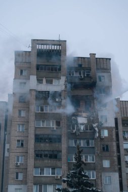 Şiddetli yangın hasarı ve yükselen dumanla büyük bir bina görüntüsü. İtfaiyeciler parçalanmış balkonlara su püskürtüyor ve gri kış gökyüzünün altında harabeler var..
