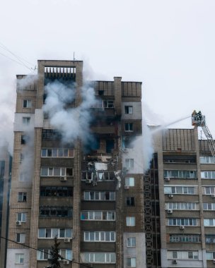 Yükseltilmiş bir Magirus döner merdivenindeki itfaiyeciler yüksek basınçlı bir hortumla patlama veya yangın sonucu hasar görmüş çok katlı bir binaya su sıkarlar. Birkaç yıkılmış balkondan yükselen duman ve kırılmış camlar görülüyor.