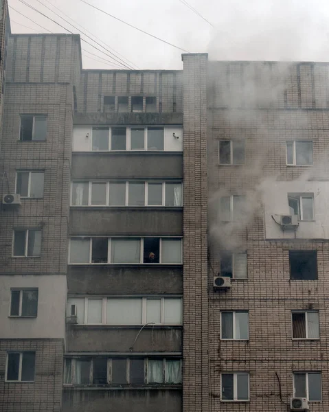 Üst katlardaki birkaç pencereden duman çıkan çok katlı gri tuğlalı bir apartmanın geniş bir görüntüsü. Duman ön cephenin hasarlı bir kısmına yoğunlaşır, ağır, bulutlu gri gökyüzüne karşı yükselir.