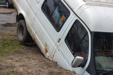 Yolda derin bir kraterde sıkışmış bir minibüs. Minibüsün ön ucu görünür durumda. Yol yüzeyinde ve yerde ciddi hasar var. Toprak hasarı ve sıkışmış aracın detayları..