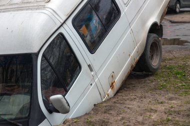 Yolda derin bir kraterde sıkışmış bir minibüs. Minibüsün ön ucu görünür durumda. Yol yüzeyinde ve yerde ciddi hasar var. Toprak hasarı ve sıkışmış aracın detayları..